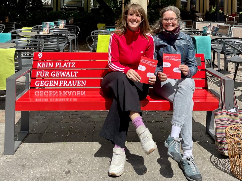 Zwei Mitarbeiterinnen der Beratungsstelle 'Beratung und Therapie für Frauen' sitzen auf der Roten Bank und lachen in die Kamera, während sie einen Infoflyer in die Kamera halten.